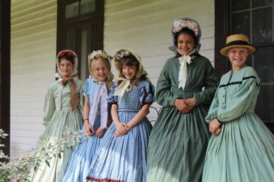 Five children stand on a porch and are dressed in 19th century women's clothing.