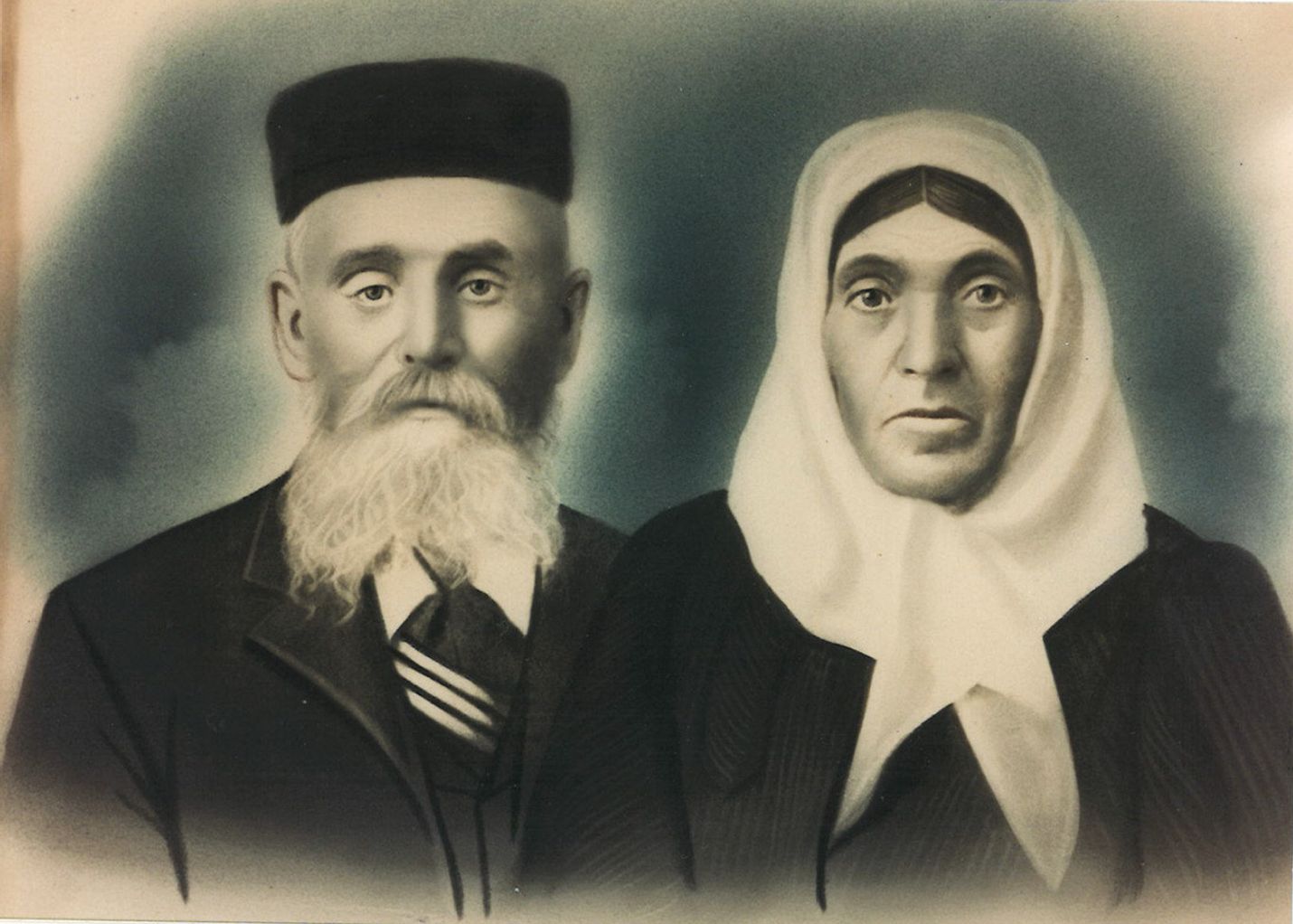 A historic photo from the late 1800s shows a man with a boxy black hat and white beard and a woman with a white scarf wrapped around her head.