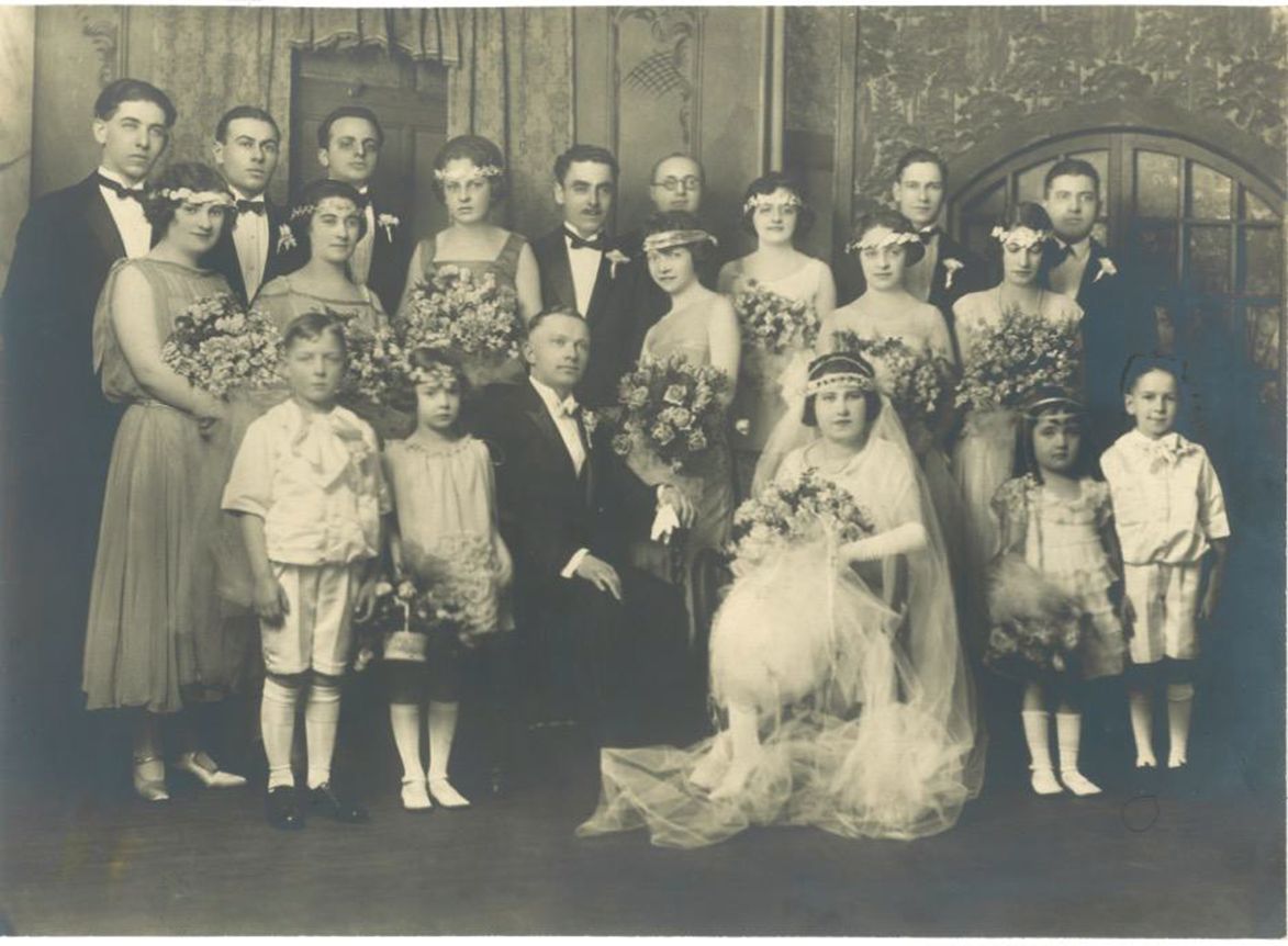 A large wedding party is pictured, with several men wearing tuxes, women holding large bouquets, and children holding baskets of flowers.