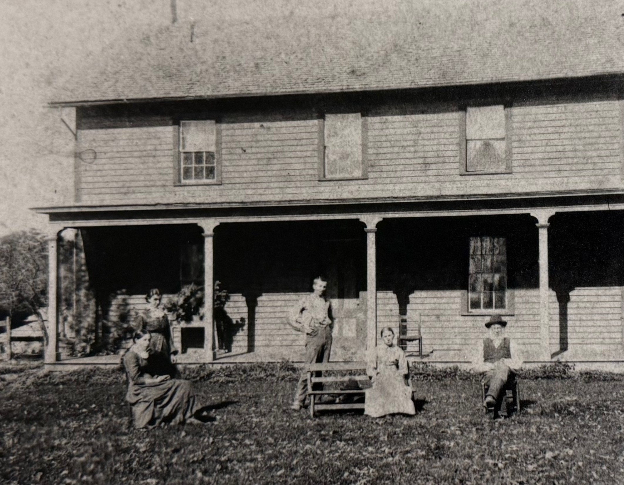 In a historical black and white photo from 1890, a family of five is standing and sitting in chairs in front of their farmhouse.