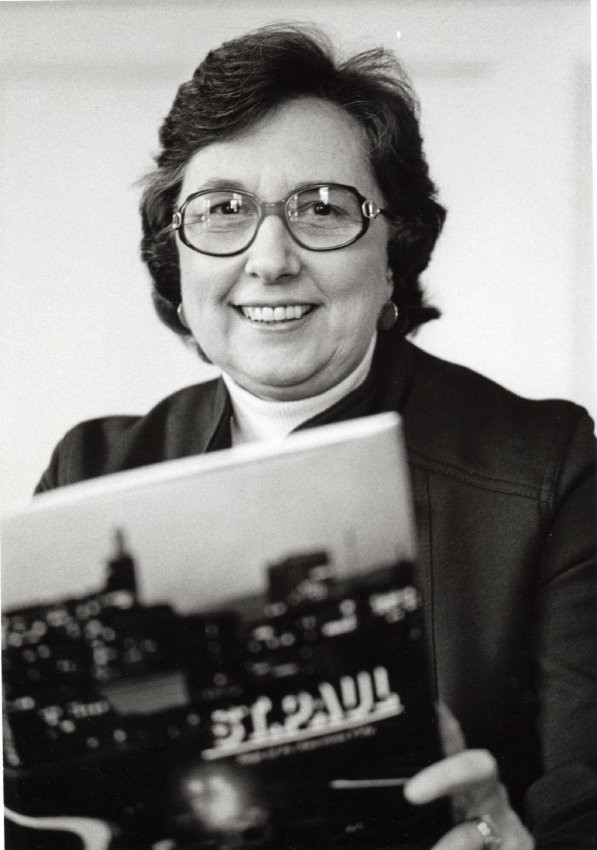 A black and white photo depicts a woman with short dark hair and large eyeglasses holding a book that says "St. Paul."